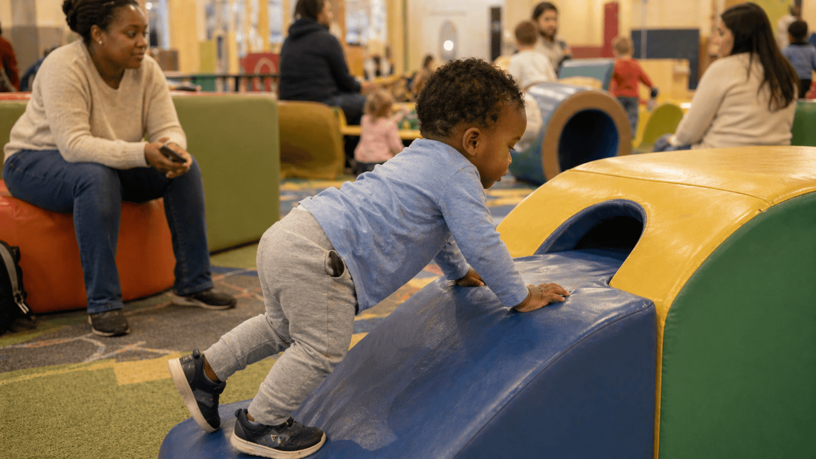 Toddler climbing at an indoor play area while parent watches nearby on a rainy day