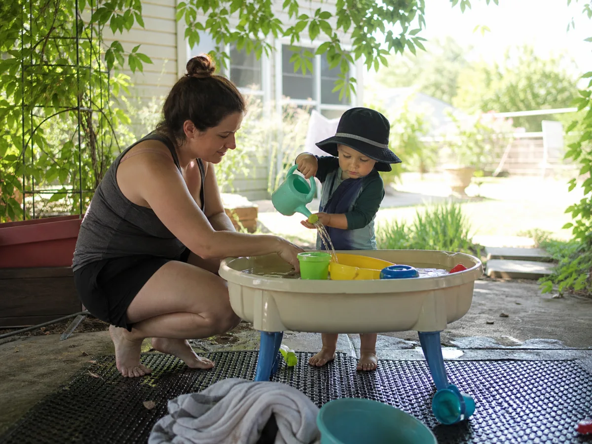 Parent supervising toddler water play under deep backyard shade