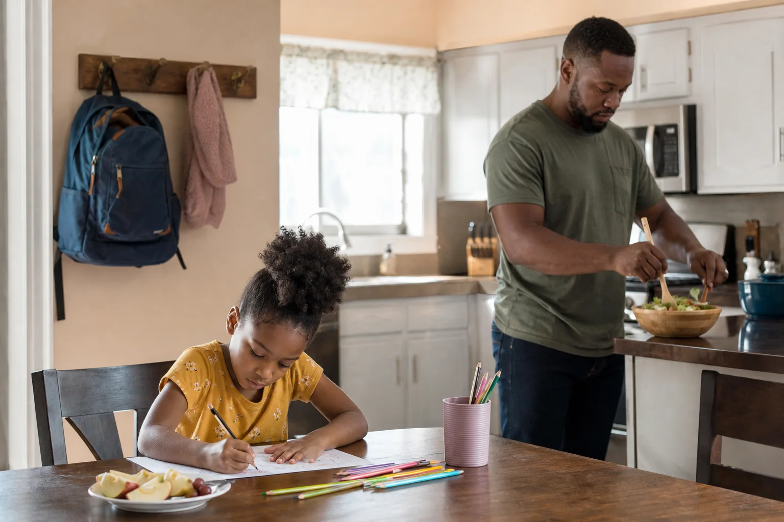 Black American father preparing dinner while his school-age daughter draws at the nearby kitchen table