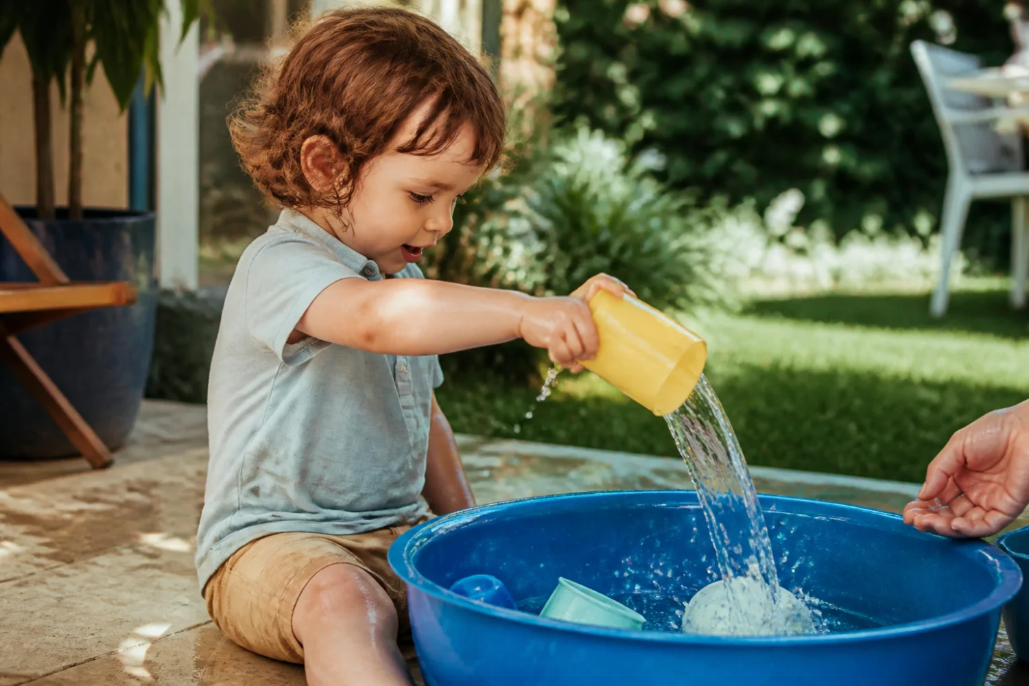 Toddler pouring water into a shallow blue tub during supervised backyard play