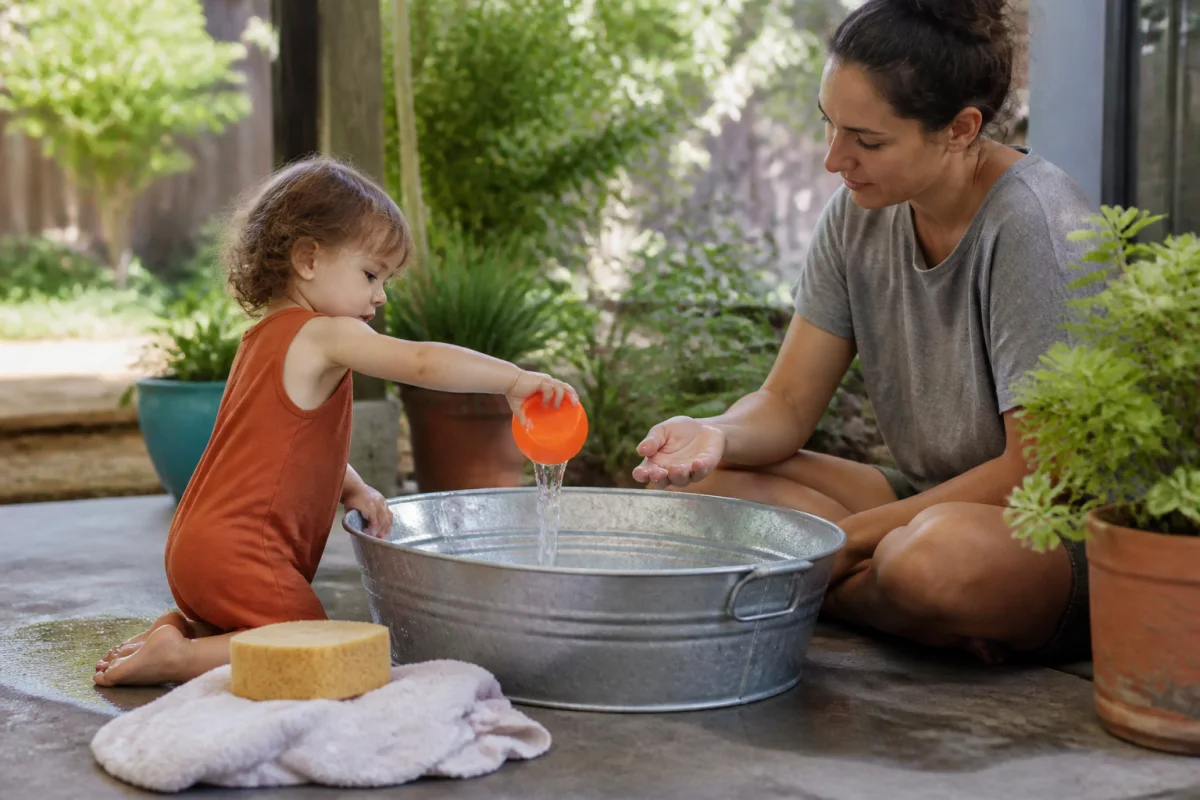 Parent sitting within arm's reach while a toddler pours water into a shallow tub
